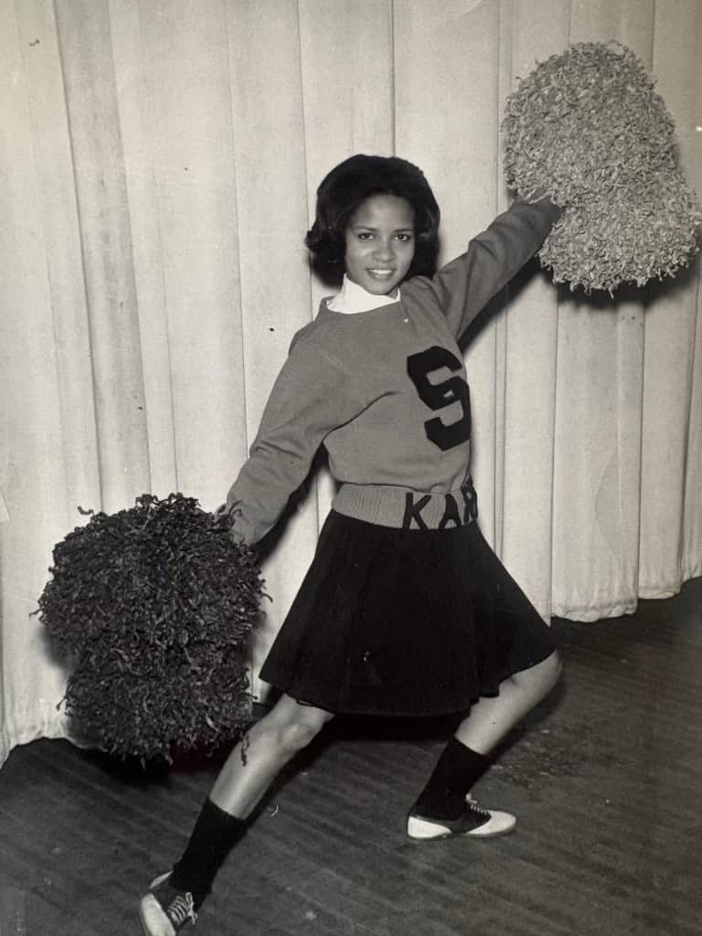Karen as a young cheerleader mid-cheer, one pom-pom raised high, her name stitched on her skirt